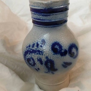 Op de afbeelding: Een witte keramische kan met blauwe en witte bloemenmotieven. De kan heeft een rond lichaam en een brede opening.