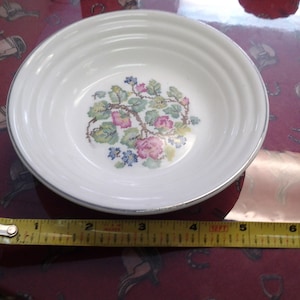 May include: A white ceramic bowl with a floral design in pink, blue, and green. The bowl has a silver rim and is approximately 5 inches in diameter.