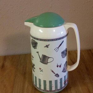 May include: A white and green insulated pitcher with a gardening theme. The pitcher has a green lid and a handle. The design features a watering can, gardening tools, and flowers.
