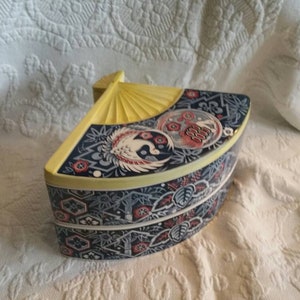 May include: A decorative box shaped like a fan with a blue, yellow and red floral design. The box is made of ceramic and has a hinged lid.