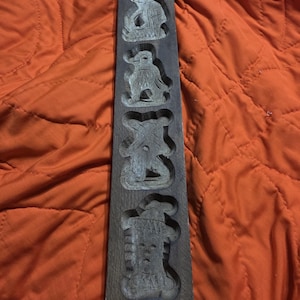 Puede incluir: Un molde de galletas de madera oscura con cuatro diseños tallados: una persona, un pájaro, un oso y una persona. El molde es rectangular y tiene un aspecto rústico. El fondo es de tela naranja.