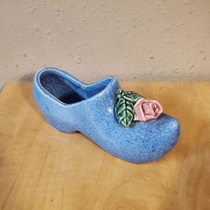 May include: A blue ceramic planter shaped like a clog with a pink rose and green leaves on the top.