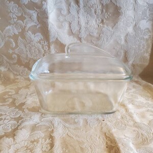 May include: A clear glass loaf pan with a lid and a handle. The pan is empty and sitting on a white tablecloth.