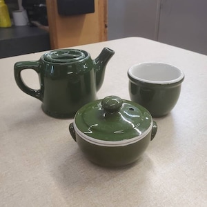 Könnte beinhalten: Eine grüne Teekanne, eine Milchkännchen und eine Zuckerdose aus Keramik. Die Teekanne hat einen weißen Rand und einen Ausguss. Die Milchkännchen und die Zuckerdose haben weiße Ränder und Deckel.