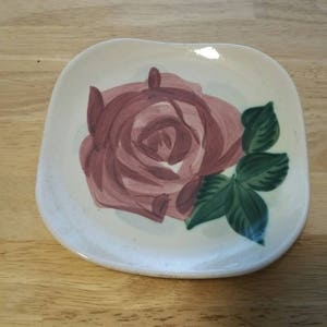 Puede incluir: Un plato de cerámica blanco con una rosa rosa y hojas verdes pintadas.
