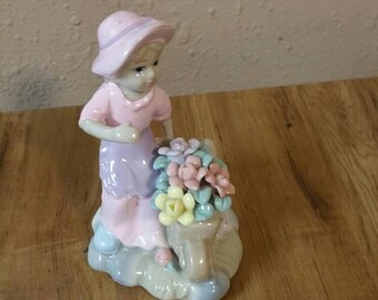 Op de afbeelding: Een porseleinen beeldje van een meisje in een roze jurk en hoed, dat een mandje met kleurrijke bloemen vasthoudt.