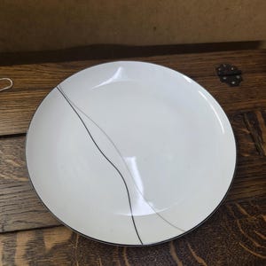 May include: A white ceramic plate with a black and gray wave design. The plate has a silver rim and is set against a wooden surface. This plate is suitable for serving meals.
