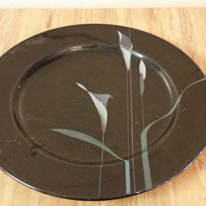 May include: A black ceramic charger plate with a stylized floral design in shades of gray and white.