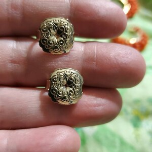 May include: A pair of gold-tone earrings with a detailed floral design. The earrings are square-shaped and have a raised, intricate pattern.