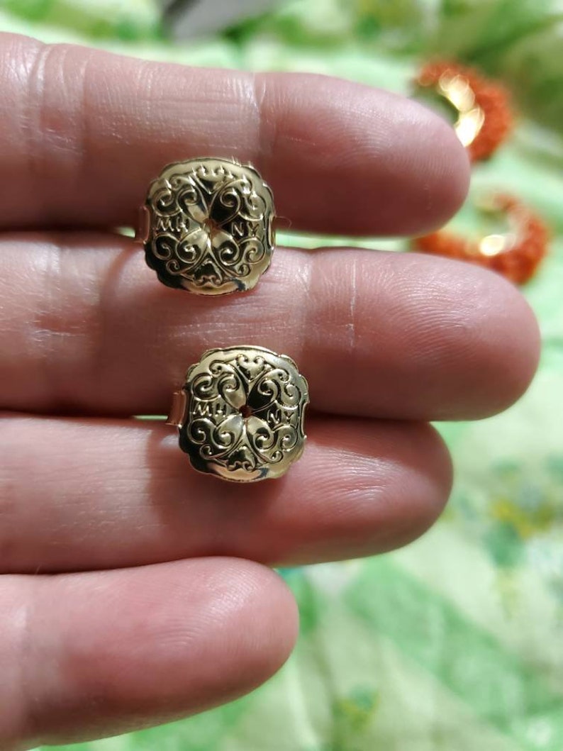 May include: A pair of gold-tone earrings with a detailed floral design. The earrings are square-shaped and have a raised, intricate pattern.