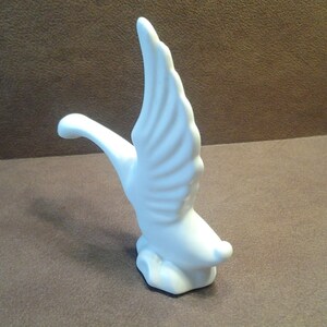 May include: A white ceramic figurine of a swan in flight. The swan has outstretched wings and a curved neck, with detailed feathering. The sculpture is set against a brown background.