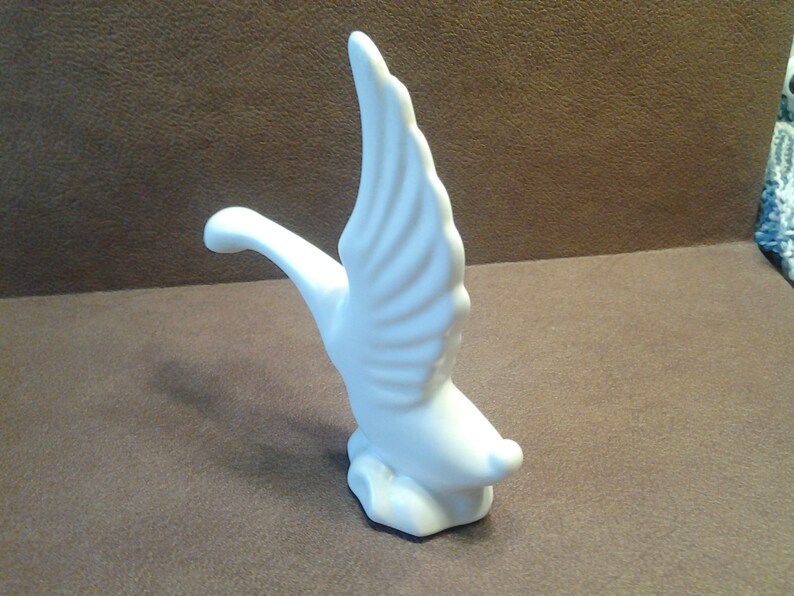 May include: A white ceramic figurine of a swan in flight. The swan has outstretched wings and a curved neck, with detailed feathering. The sculpture is set against a brown background.
