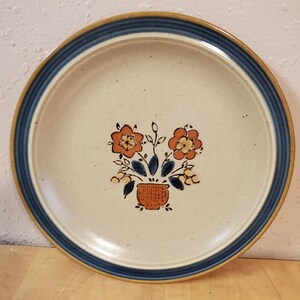 Op de afbeelding: Een beige keramisch dinerbord met een blauwe en bruine rand. Het bord heeft een handgeschilderd ontwerp met oranje bloemen in een bruine pot met groene bladeren.