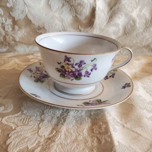 May include: A white teacup and saucer set with a delicate floral pattern of purple and yellow flowers. The set has a gold trim around the edges.