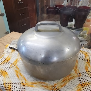 May include: A round, silver-colored aluminum Dutch oven pot with a matching lid. The pot features two side handles with a ribbed design and a central handle on the lid. The metal surface shows signs of wear and minor dark spots throughout the exterior.
