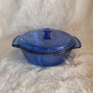 Puede incluir: Un plato para hornear de vidrio azul con tapa. El plato tiene dos asas a los lados y una perilla en la tapa.
