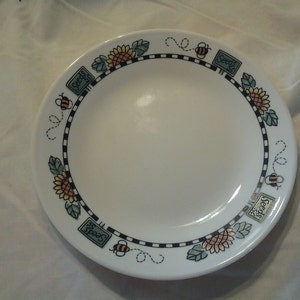 May include: White ceramic plate with a decorative border featuring sunflowers, bees, and the word "Seeds" in a rectangular frame. The border is black and white striped with a dotted line.