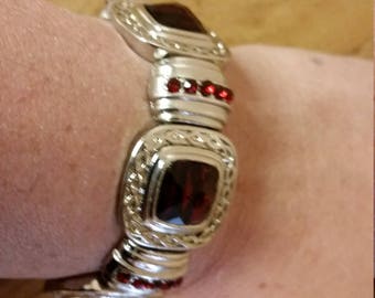 May include: Silver bracelet with red gemstone accents. The bracelet is made of metal and has a square shape. The gemstones are set in the metal and are surrounded by smaller stones.
