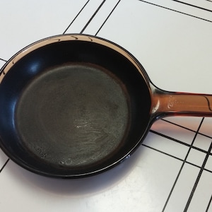 May include: A black skillet with a brown handle. The pan is round with a dark interior and a clear, amber-colored handle. The handle has a curved design. The skillet is on a white surface with black lines.