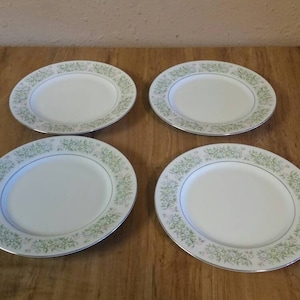 Puede incluir: Juego de cuatro platos de cena de cerámica blanca con un patrón floral verde. Los platos tienen un borde ligeramente elevado y un acabado liso y brillante.