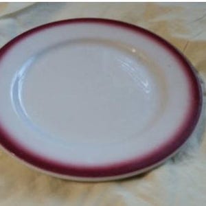 Puede incluir: Un plato de cerámica blanco con un borde granate. El plato tiene un diseño simple y clásico, adecuado para servir alimentos. El plato es redondo y parece estar en buenas condiciones.