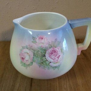 May include: A white ceramic jug with a pastel blue, pink and yellow ombre glaze. The jug features a floral design with pink roses and green leaves.