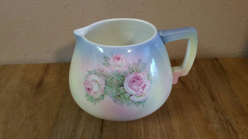 May include: A white ceramic jug with a pastel blue, pink and yellow ombre glaze. The jug features a floral design with pink roses and green leaves.