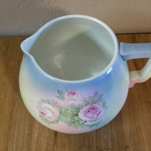 May include: A vintage ceramic pitcher with a light blue, pink and yellow ombre glaze. The pitcher features a floral design with pink roses and green leaves.