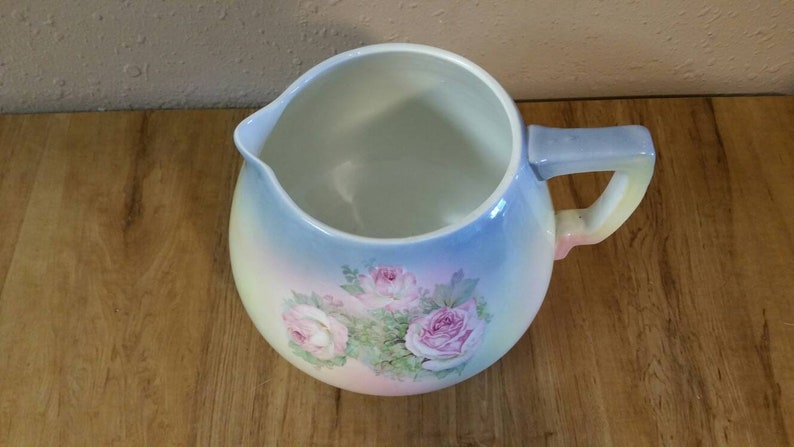 May include: A vintage ceramic pitcher with a light blue, pink and yellow ombre glaze. The pitcher features a floral design with pink roses and green leaves.