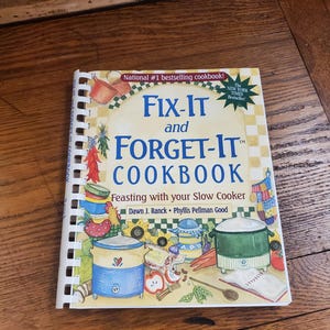 May include: A cookbook titled "Fix-It and Forget-It Cookbook" with a colorful illustration of a slow cooker, vegetables, and other kitchen items. The book is a New York Times Bestseller and features recipes for slow cooker meals.