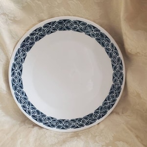 May include: A white ceramic dinner plate with a blue and white patterned border. The pattern is a repeating design of stylised leaves and swirls.