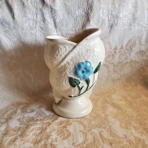 Op de afbeelding: Een witte keramische vaas met een getextureerd oppervlak en een blauw bloemenontwerp. De vaas heeft een unieke vorm, met een gebogen opening en een brede basis.