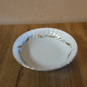 Puede incluir: Un tazón de cerámica blanca con un borde dorado y un diseño floral verde y dorado. El tazón está sobre una superficie de madera.