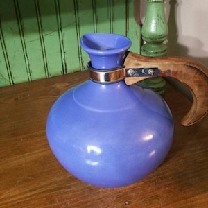 May include: A blue ceramic pitcher with a wooden handle and a silver band around the neck. The pitcher is sitting on a wooden surface.