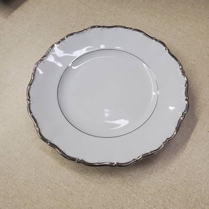 Könnte beinhalten: Ein weißer Porzellanteller mit silbernem Rand. Der Teller hat einen gewellten Rand und eine glatte, glänzende Oberfläche.