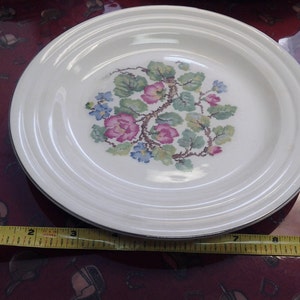 Könnte beinhalten: Ein weißer Keramikteller mit einem Blumendesign mit rosa, blauen und grünen Blumen. Der Teller hat einen erhöhten Rand und einen silbernen Rand.