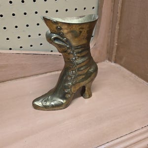 May include: A vintage brass boot-shaped vase. The boot has a pointed toe, a small heel, and is adorned with decorative buttons and detailing. The vase is displayed on a light pink surface, suggesting a decorative or antique theme.