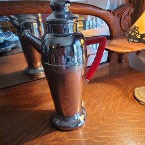 Könnte beinhalten: Ein silberner Cocktailshaker mit einem roten Kunststoffgriff. Der Shaker hat eine abgerundete Oberseite und einen zylindrischen Körper. Er steht auf einer Holzoberfläche.