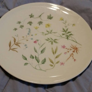 May include: A white ceramic plate with a floral design featuring green leaves, pink and yellow flowers. The plate has a slightly raised rim.