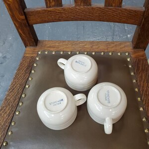 以下が含まれることがあります： 底に「Stoneware Japan」と青い文字が書かれた白いセラミック製のマグカップが3つ。マグカップは茶色の革の表面の上に置かれています。