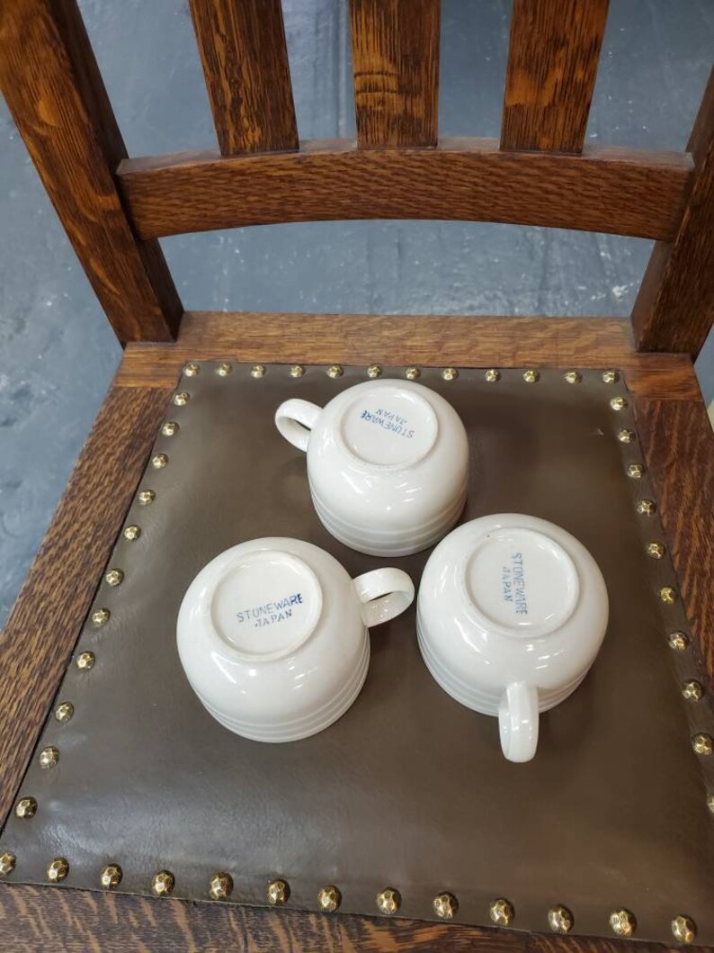 以下が含まれることがあります： 底に「Stoneware Japan」と青い文字が書かれた白いセラミック製のマグカップが3つ。マグカップは茶色の革の表面の上に置かれています。