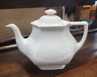 Może przedstawiać: Biały ceramiczny czajnik z pokrywką i uchwytem. Czajnik ma ozdobny wzór na korpusie.