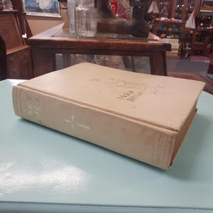 Könnte beinhalten: Ein geschlossenes, cremefarbenes, gebundenes Buch mit den Worten "Holy Bible" auf dem Rücken. Das Buch liegt auf einer hellblauen Oberfläche.