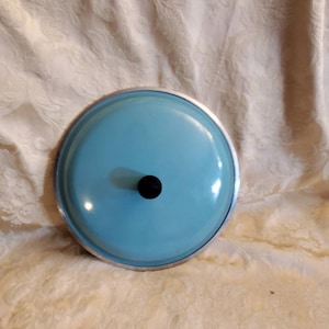 Puede incluir: Una tapa de olla de esmalte azul claro con un borde plateado y un pomo negro.