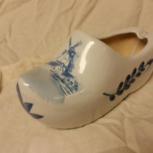 May include: A white ceramic clog with blue and white Delftware designs, featuring a windmill and floral motifs. The clog is a planter with a hollow interior.