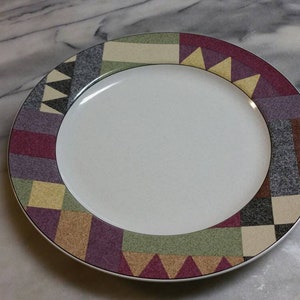 Może przedstawiać: Biały ceramiczny talerz obiadowy z kolorową geometryczną obwódką. Obwódka zawiera odcienie czerwieni, fioletu, żółci, zieleni i brązu.
