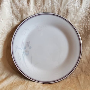 May include: A white ceramic dinner plate with a delicate floral design in shades of gray and purple. The plate has a thin gold rim and a wider burgundy rim.
