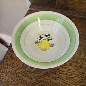 May include: A white ceramic bowl with a green and white striped rim. The bowl features a yellow lemon illustration with green leaves in the center. The bowl is likely for serving food.