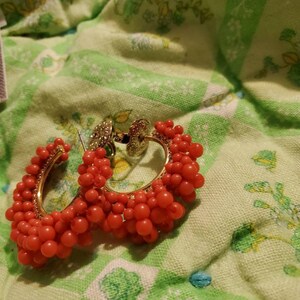 May include: A pair of hoop earrings with a gold base and red beads. The beads are clustered together to create a textured look.