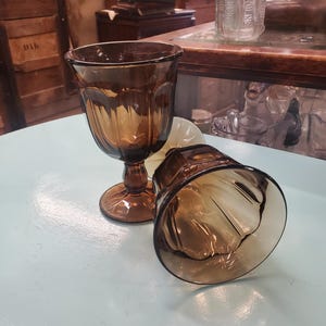 May include: Two brown glass goblets with a fluted design. The goblets are on a light blue surface.
